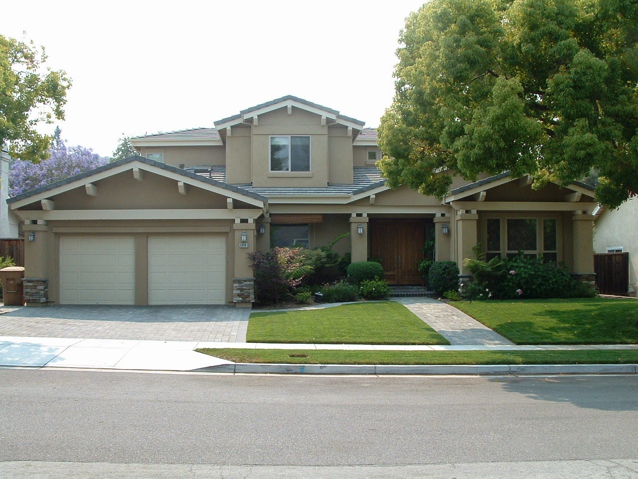 A two-story suburban house with beige exterior, a double garage, and a well-maintained front yard. The home boasts an elite gabled roof with green trim and shutters, and is partially shaded by a large tree on the right side, adding to its elevated charm.