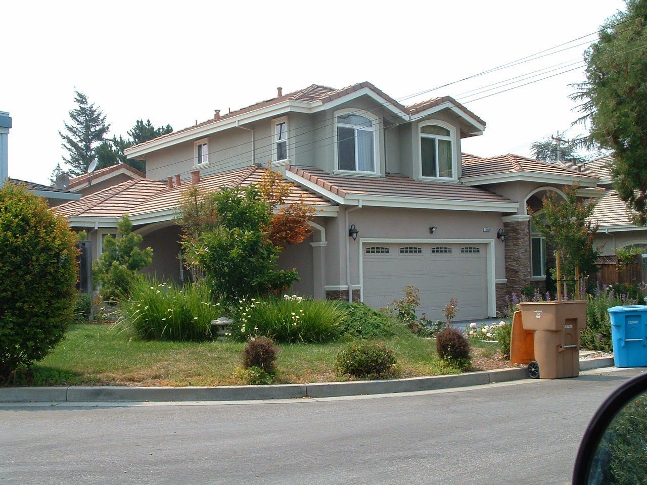 Projects 8 A two-story suburban house with beige siding and a tiled roof rests at an elite elevation. The front yard is landscaped with bushes and trees, while tan and blue trash bins sit curbside. The garage, part of the main structure, features two doors.