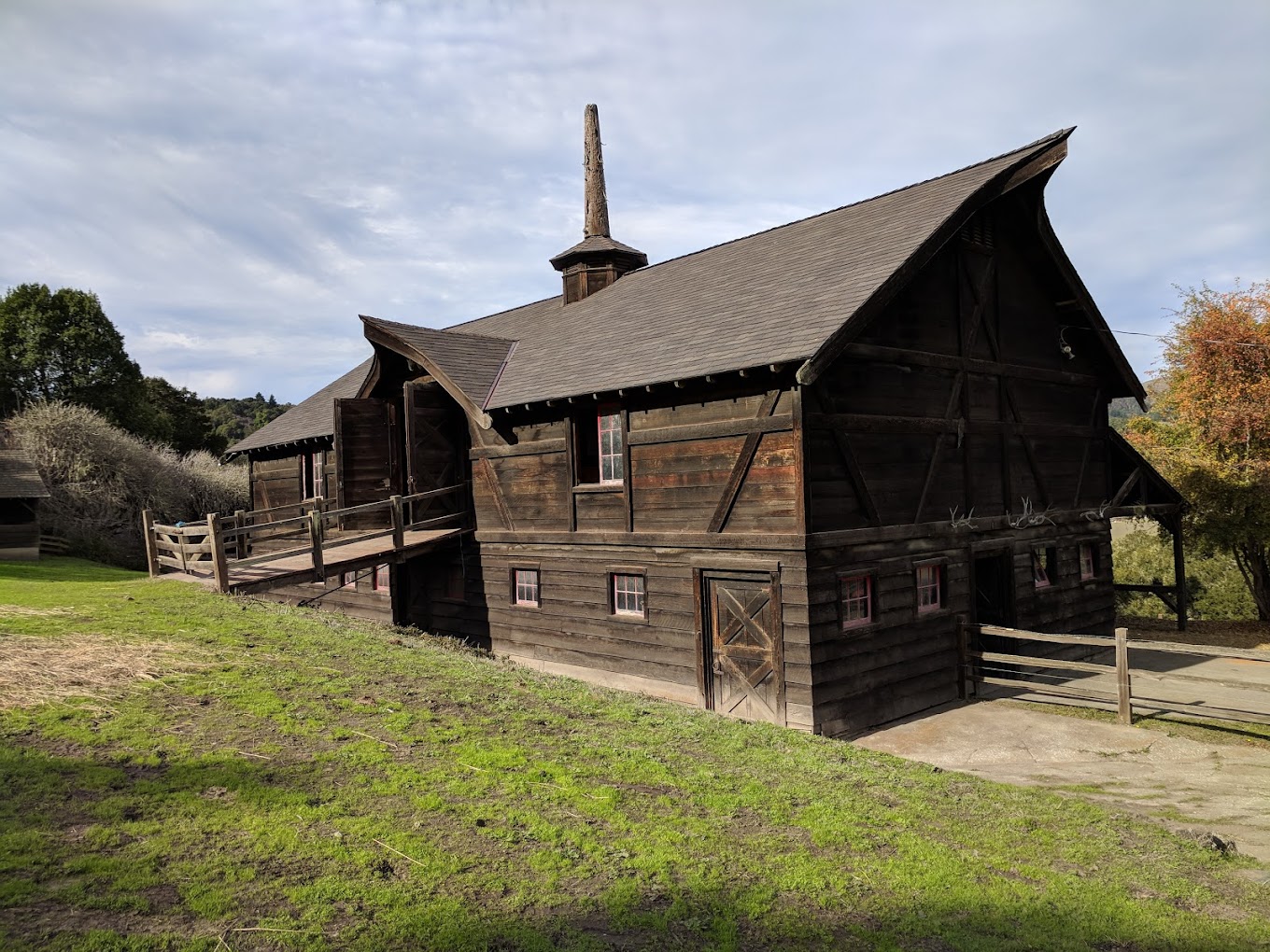 Projects 7 A rustic, wooden barn with dark brown planks stands on a grassy slope under a cloudy sky. The barn, demonstrating the fine craftsmanship of Elevation Elite's roofing services, has a steep, shingled roof with a small cupola, several small windows, and a ramp leading to a door on the upper level. Trees surround the area.