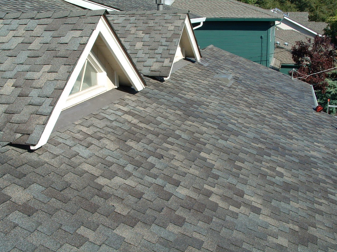 Projects 5 A close-up view of a residential roof with gray asphalt shingles, showcasing ClassicTouch. The roof features dormer windows with white trim. In the background, a neighboring house painted green is visible. Trees surround the houses under a clear sky.