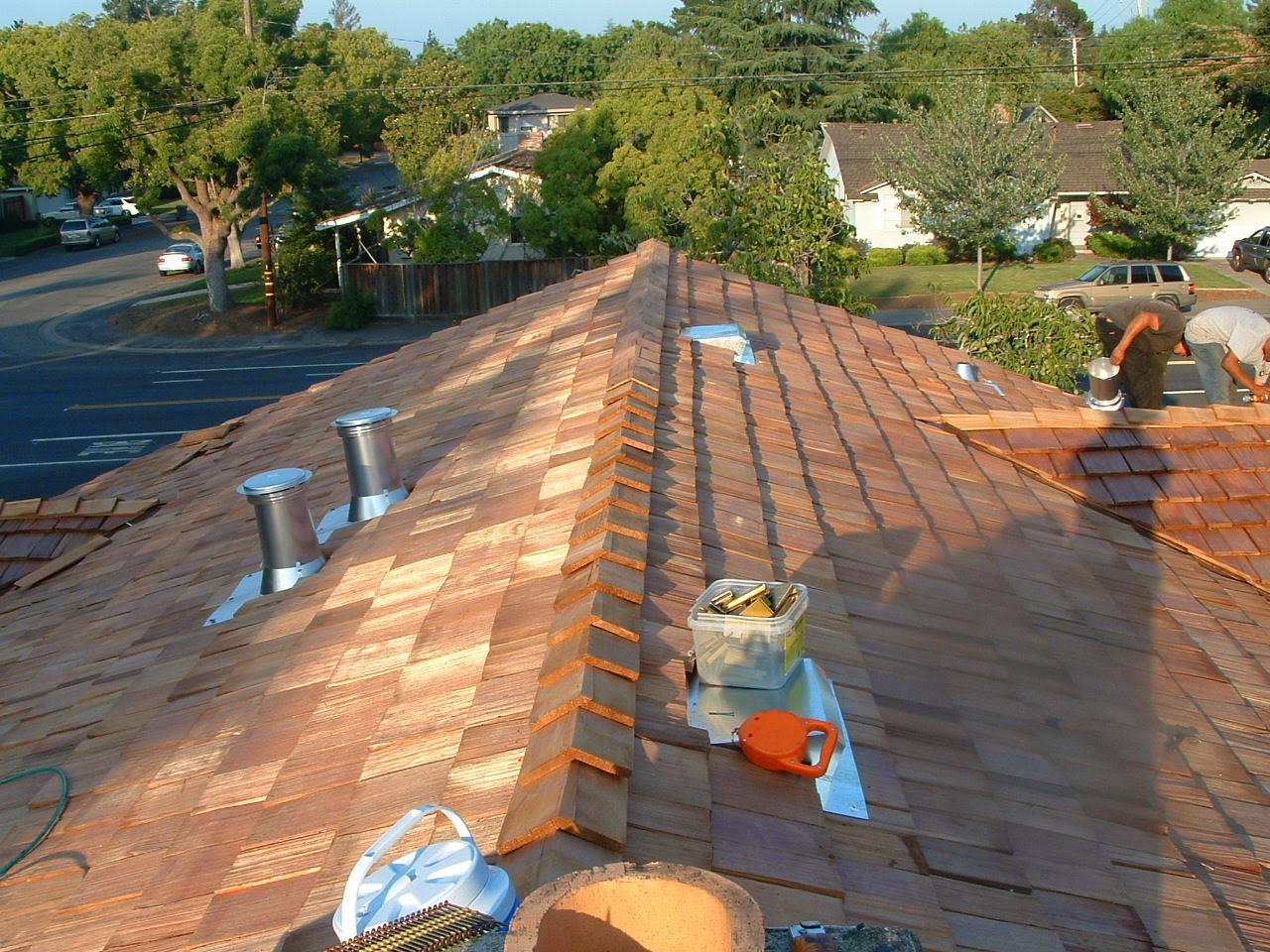 Projects 3 View from the ridge of a wooden shingle roof, showing fresh installation. Several tools and materials are spread out. Two people are working on the roof, showcasing their expertise in Shingle Renewal. In the background, houses, trees, and a street add to the picturesque scene.
