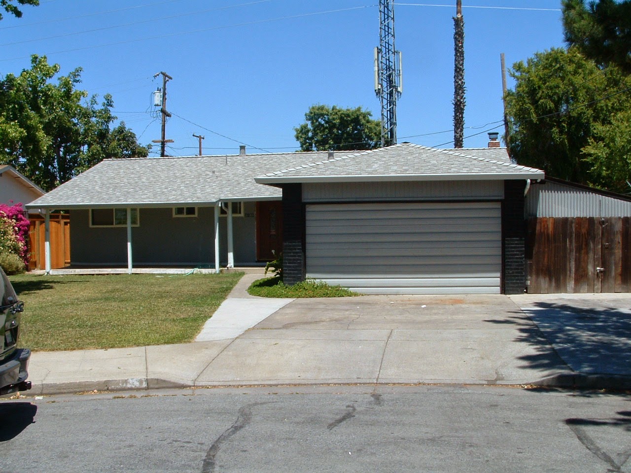 Projects 4 A single-story house with a classic gray shingle exterior and a two-car garage. The front yard has a small grassy area and a concrete driveway. There are trees in the background, a wooden fence on the right, and utility poles and a cell tower visible behind the house, adding both charm and renewal.