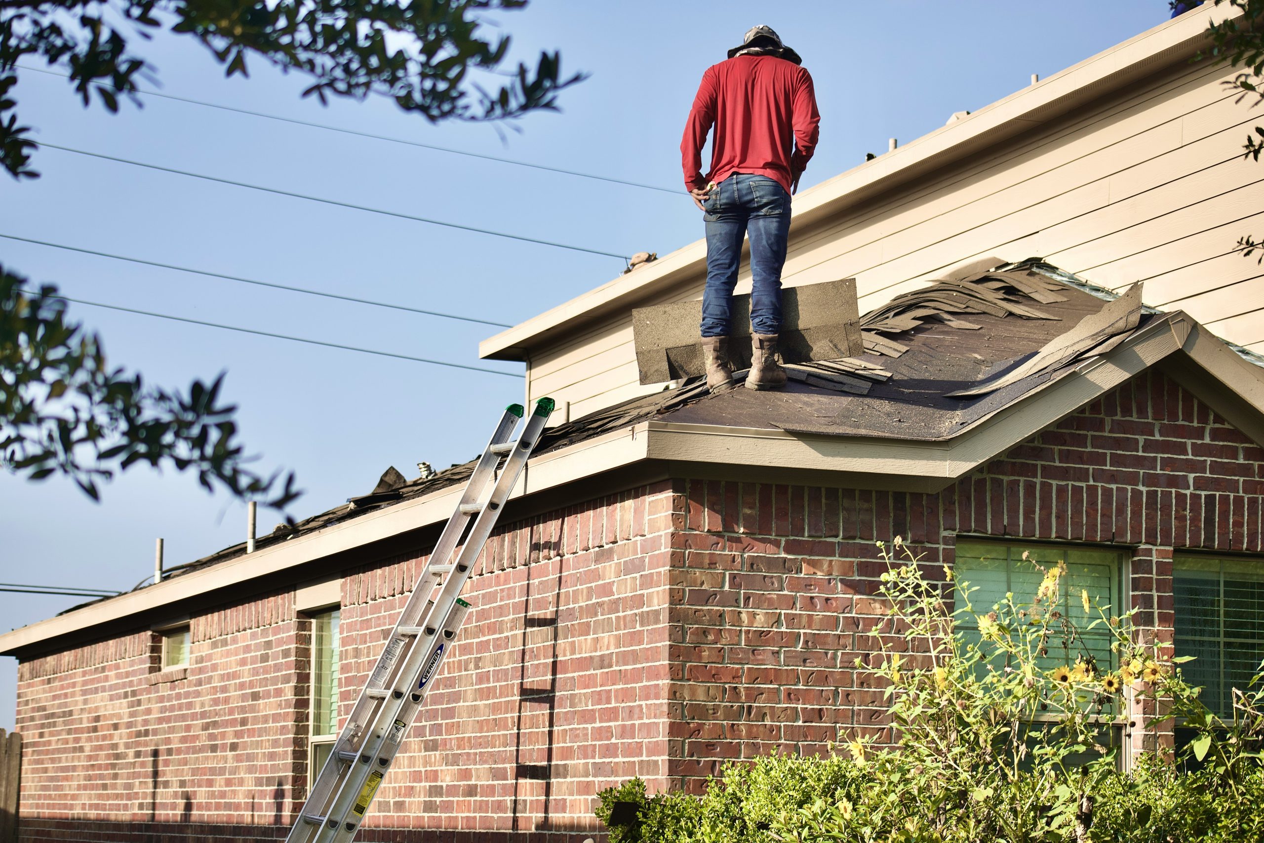 A person wearing a red hoodie and jeans stands on the roof of a brick house, performing maintenance or repairs likely related to storm damage. A long metal ladder is propped against the side of the house, leading up to the roof. The sky is clear and blue, perfect for getting a roof replacement cost estimate.