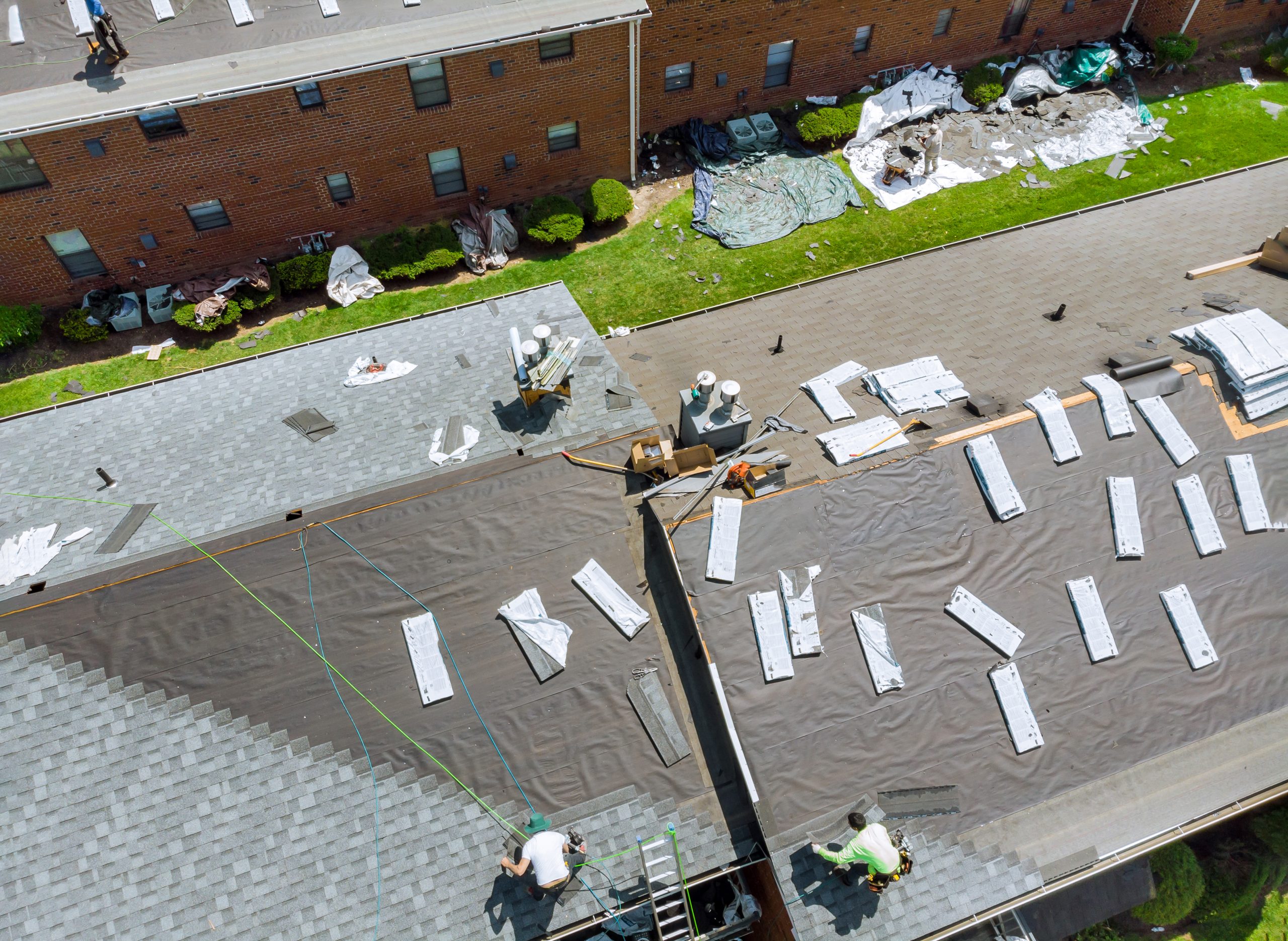Bird's-eye view of roofers working on a residential building. The roof is partially covered with new shingles, suggesting a roof replacement in progress. Roofing materials are scattered around, and the roofers are actively working with visible equipment and tools. Surrounding area includes brick buildings and greenery.