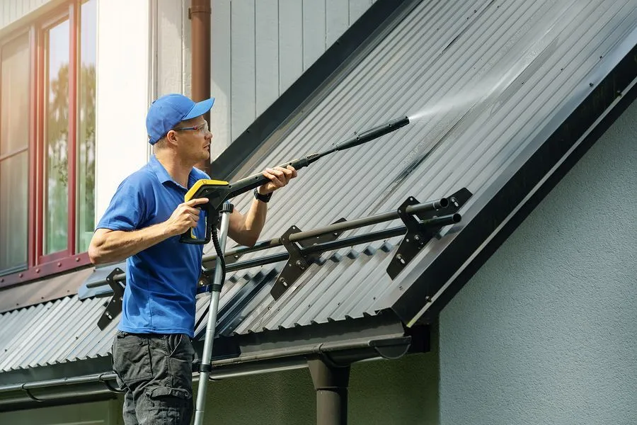 A person wearing a blue cap and shirt uses a pressure washer for roof maintenance, cleaning a metal roof. The individual is standing next to a light gray building with a red window frame. Sunlight streams in from the left side, illuminating part of the scene.