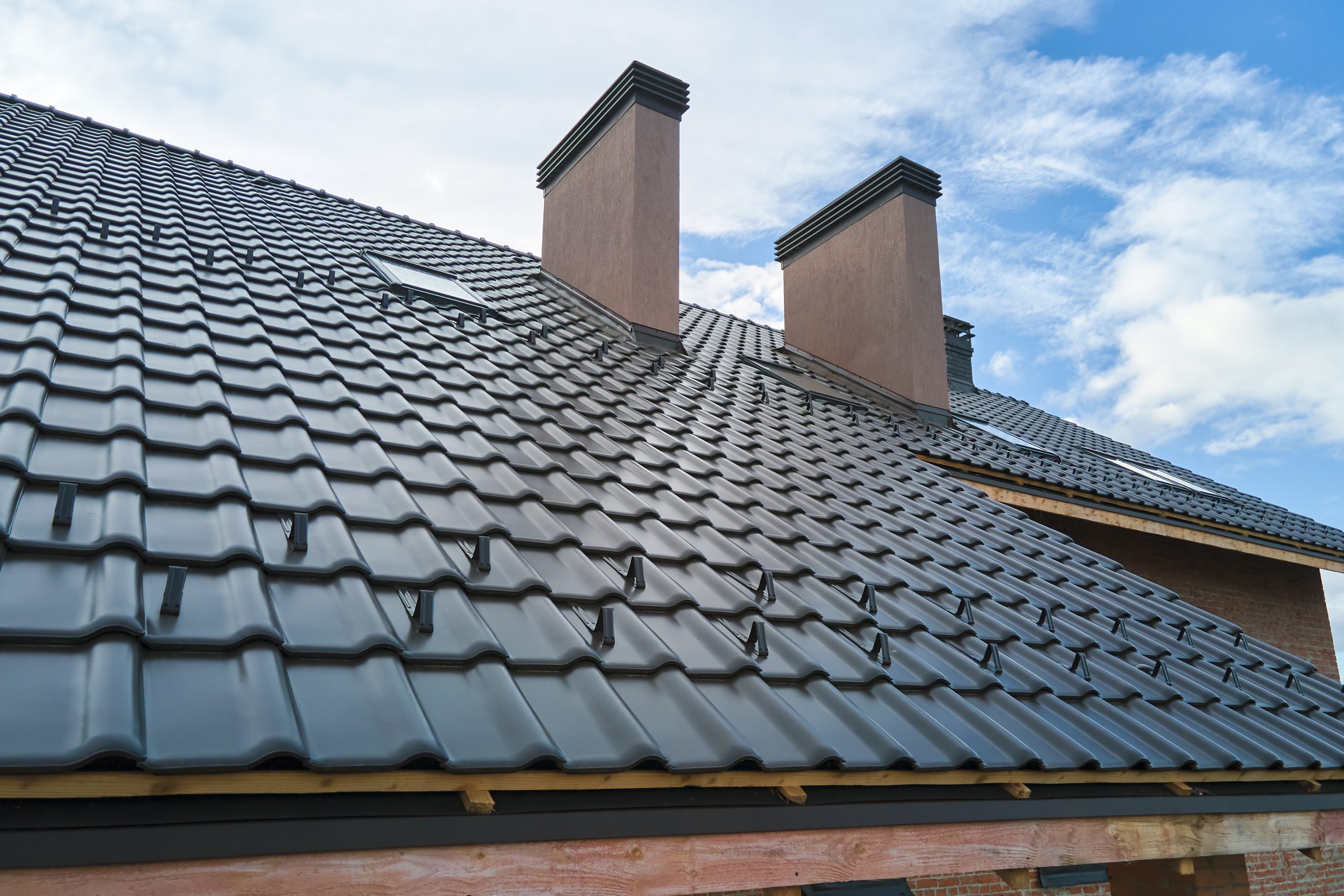 A close-up view of a house roof with black ceramic tiles, featuring two chimneys and a small skylight. The self-cleaning roof shines under the bright sky with scattered clouds. Wooden planks line the edges, adding a touch of rustic charm.