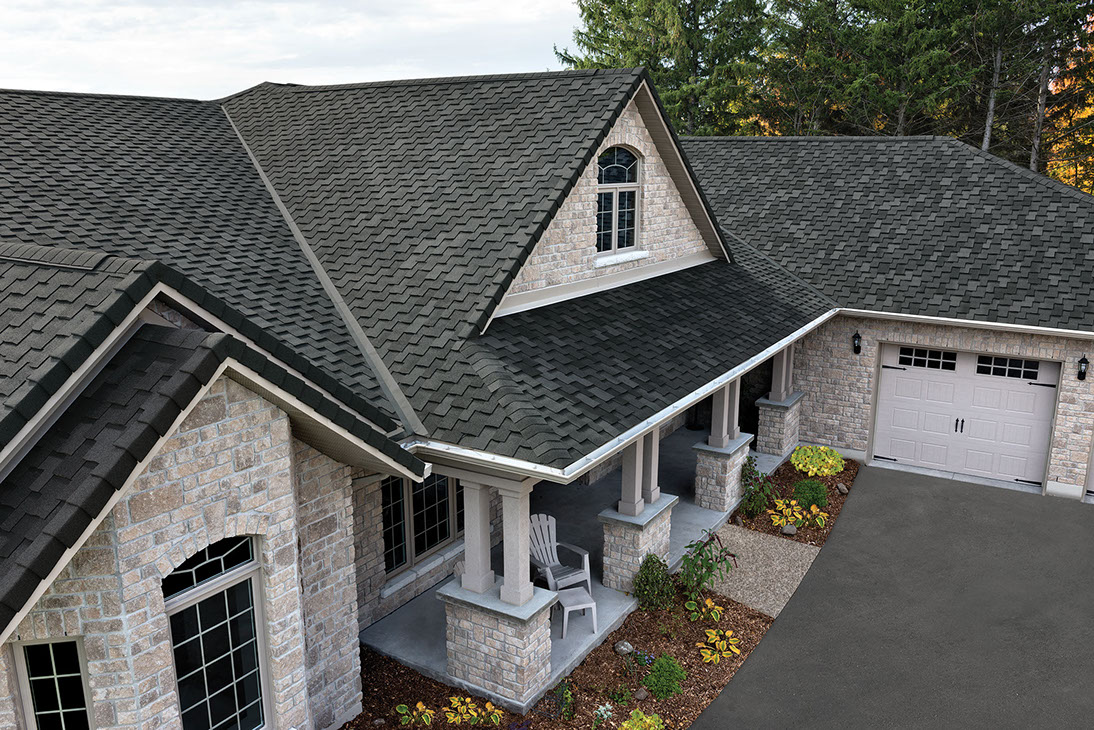 A single-story house with light-colored brick walls and a dark, asphalt shingle roof. It has an arched window under the gable and two gray chairs on a covered front porch with columns. The driveway leads to a two-car garage. Small plants surround the entrance, enhancing the home's welcoming appeal.
