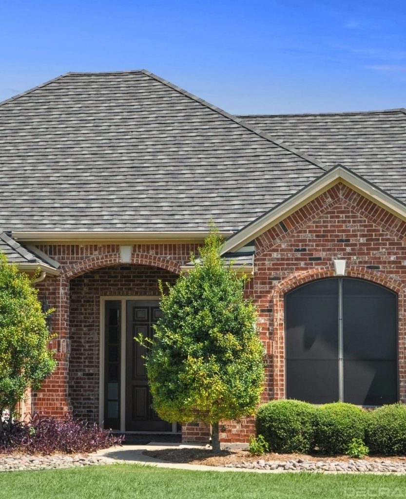 A brick house with a dark shingled roof features a small porch with an arched entrance supported by two columns. The front yard is landscaped with a mix of shrubs, small trees, and a neatly trimmed lawn against the backdrop of a clear blue sky, perfectly complementing the elegant shingle roofing.