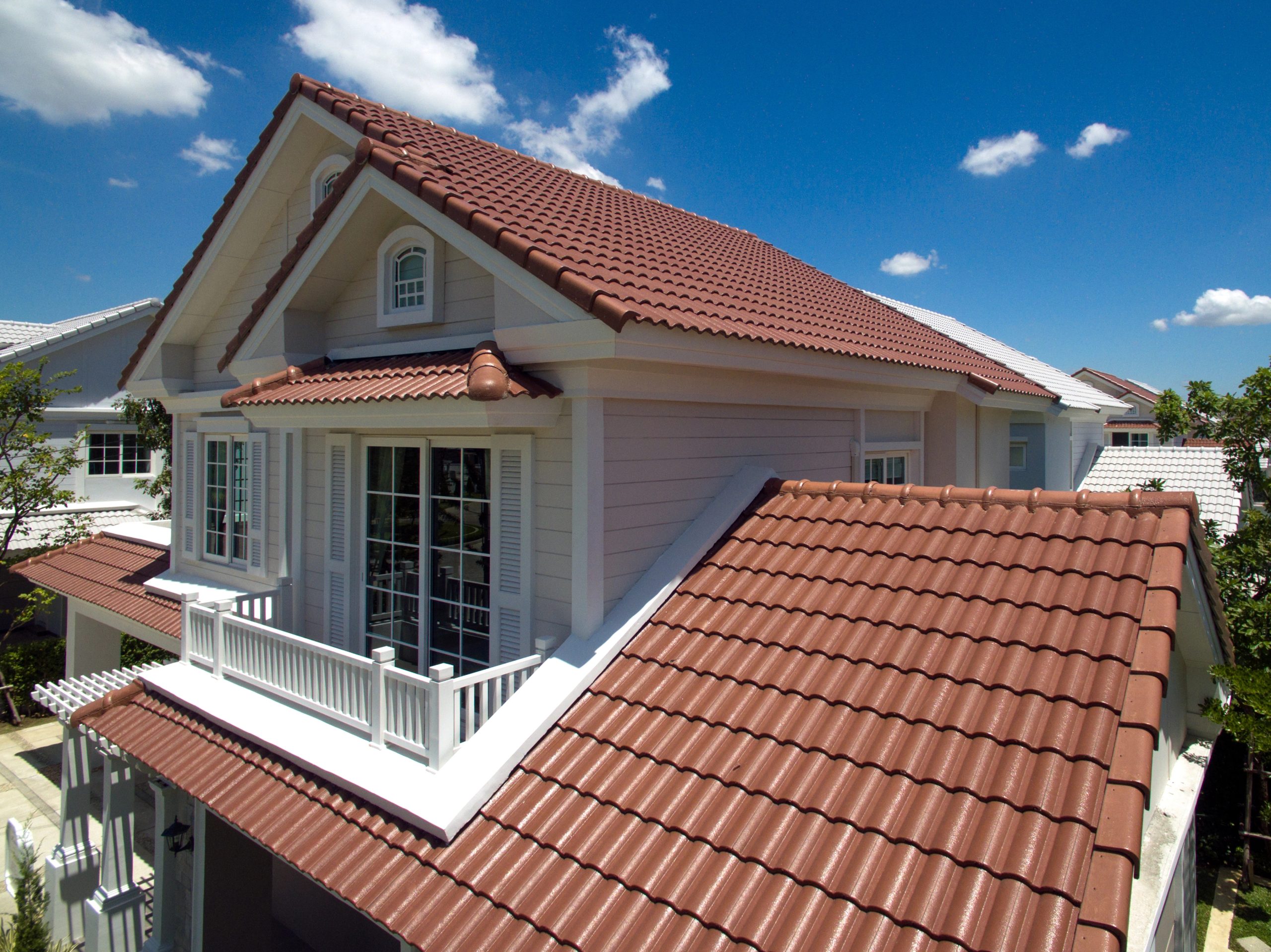 A modern, two-story house with a red tile roof, light-colored exterior walls, and large windows. The upper floor has a balcony with white railing and decorative trim. The sky is clear and blue with a few scattered clouds, and there are trees surrounding the house—showcasing the Tile Roof Benefits.