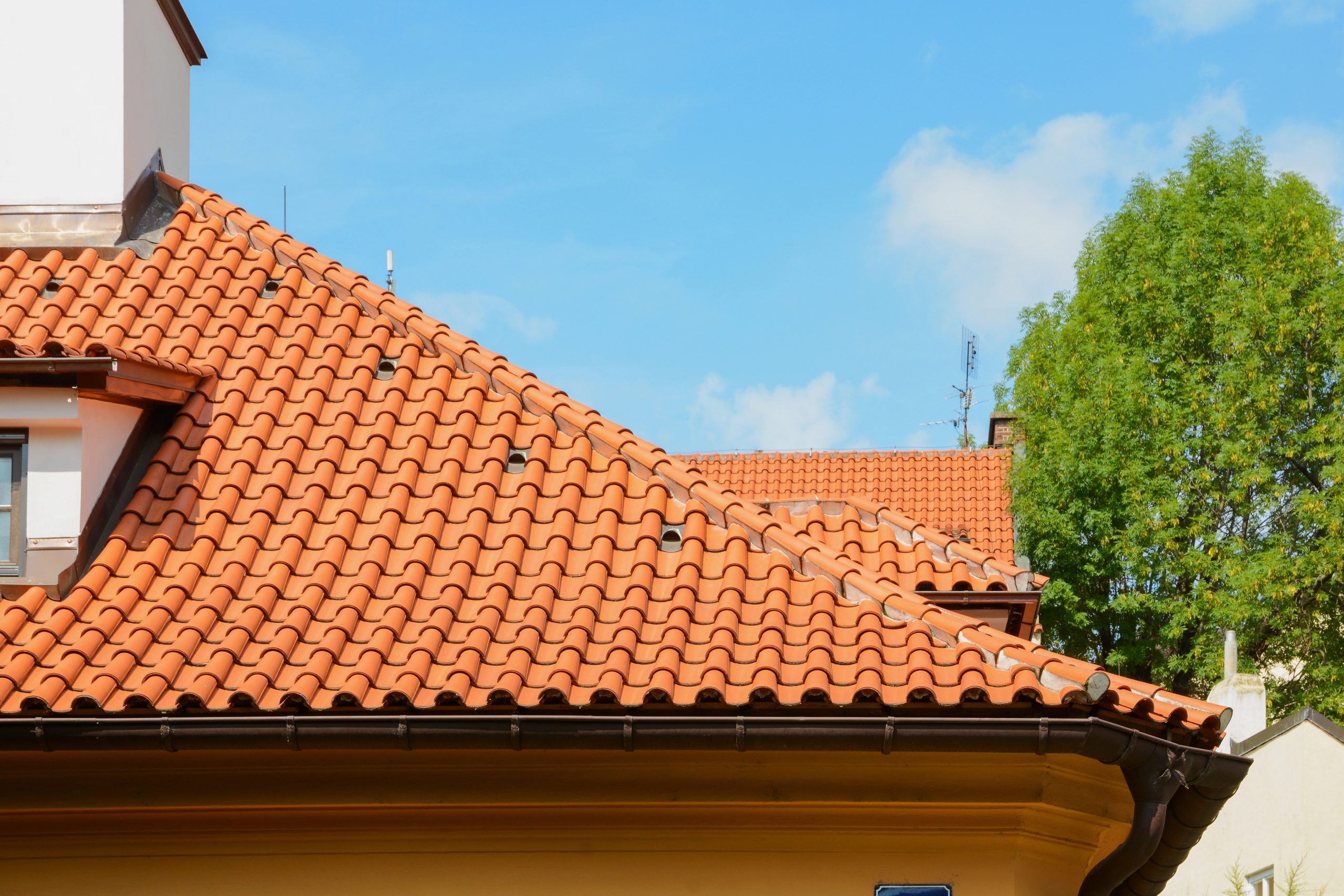 A close-up of buildings with traditional red-tiled roofs on a sunny day. The rooftops feature classic Ludowici clay tiles, and a portion of a tree is visible in the background under a clear blue sky.