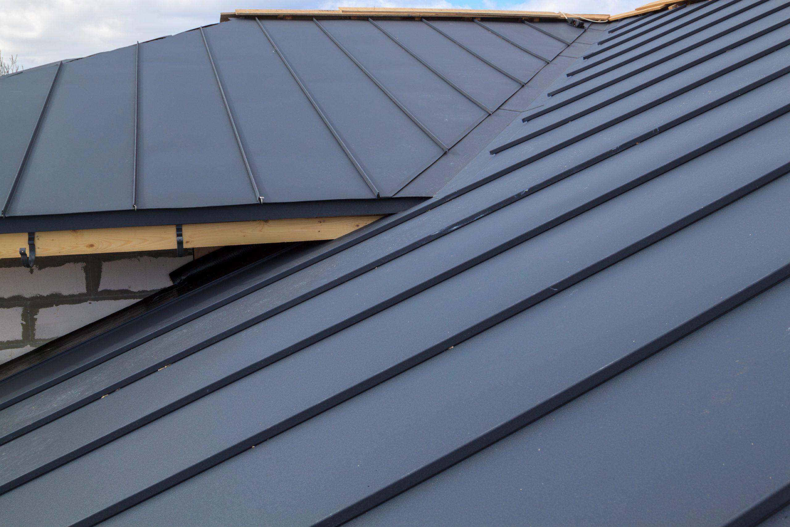 A close-up of a dark grey metal roof with a modern, sleek finish. The roof panels are arranged in a standing seam pattern. Part of an unfinished brick structure can be seen beneath the roof edge, with some light wood framing visible. The sky is slightly cloudy.