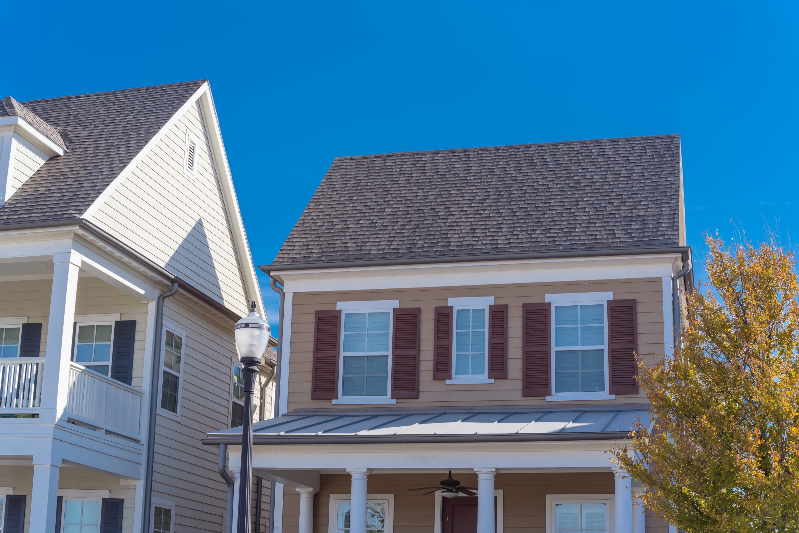 A two-story house with beige siding, white trim, and dark brown shutters stands on a sunny day. It features a covered porch with white columns and composite roof shingles. A street lamp is visible in the foreground, with a partly visible neighboring house on the left.