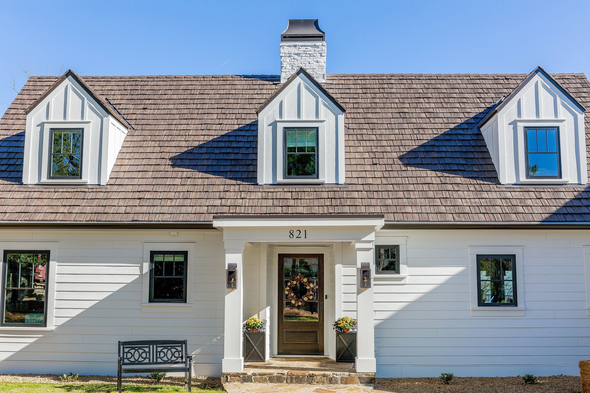 A quaint, white, two-story house with a brown composite shingle roof, three dormer windows on the upper floor, and a central brick chimney. The front entrance features a wooden door with glass panes and the house number "821" above it. A small black bench sits to the left.