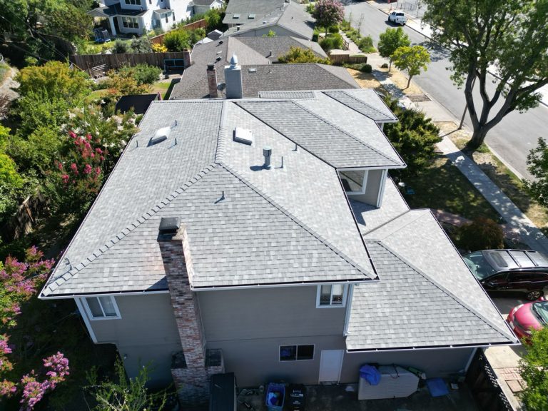 Aerial view of a residential neighborhood showcasing a large, two-story house with a gray shingled roof and brick chimney, featuring recent Roofing Installation. Surrounding houses with similar roofs are visible, along with trees, a street lined with cars, and a sidewalk bordering the area.
