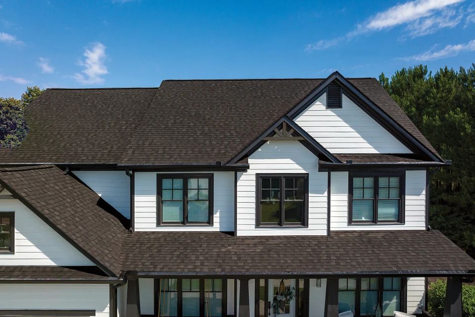 A modern two-story house with white siding and dark shingles on the roof. The house features multiple large windows, a peak roof design, and black trim accents. The roof is adorned with composite shingles from top synthetic shingle manufacturers. There is a small decorative gable above the central window on the upper floor, all set against a bright blue sky background.