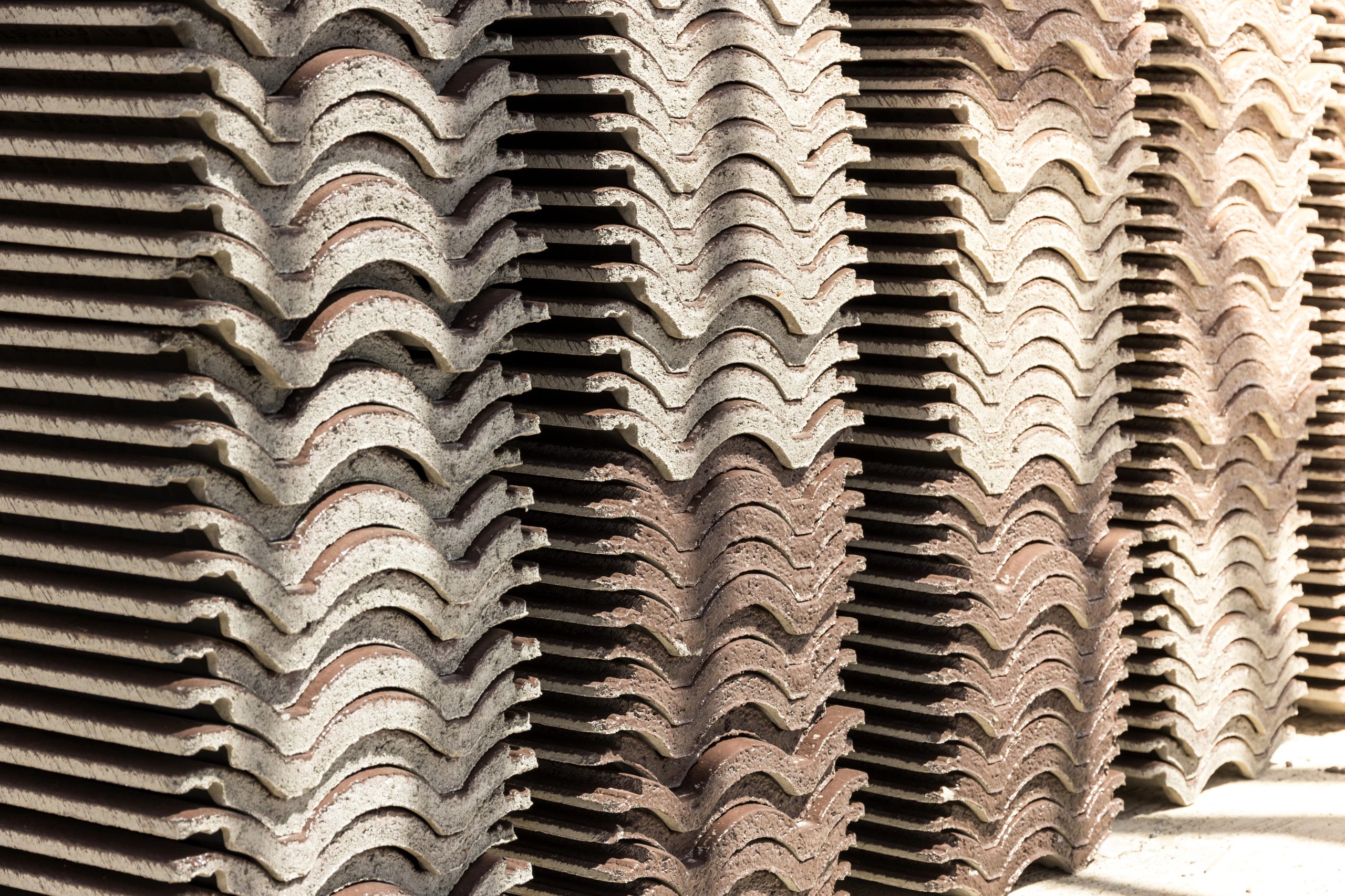 Stacks of brown and gray concrete tile roofing sit in a neat vertical formation, their style and strength highlighted by sunlight casting shadows to emphasize their wavy, interlocking shapes.