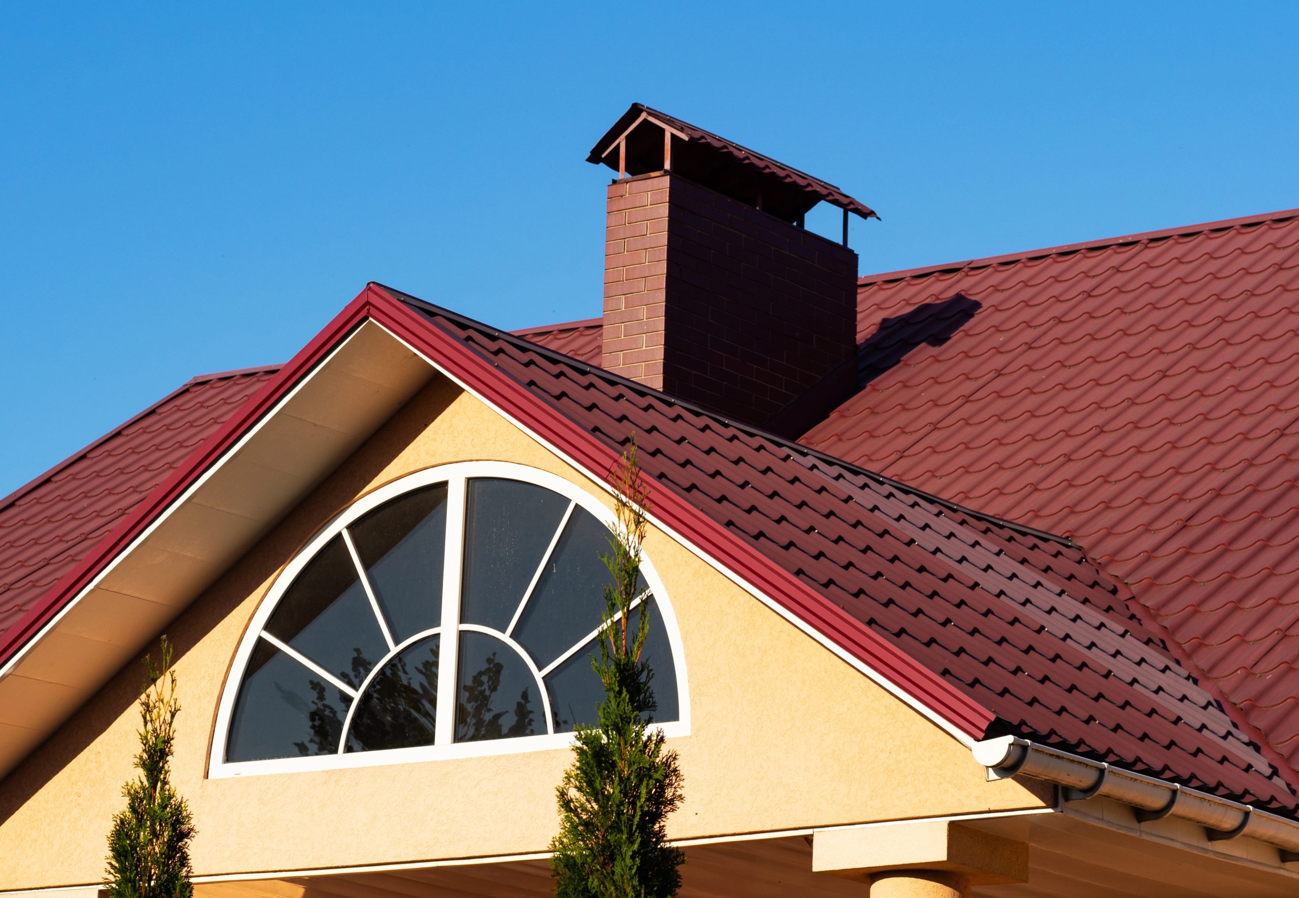 The elegant house with a yellow stucco exterior features a red metal tile roof and a brick chimney. An arched window with white trim graces the roof, while two tall, narrow evergreen trees stand in the foreground against the clear blue sky, adding strength to its picturesque setting.