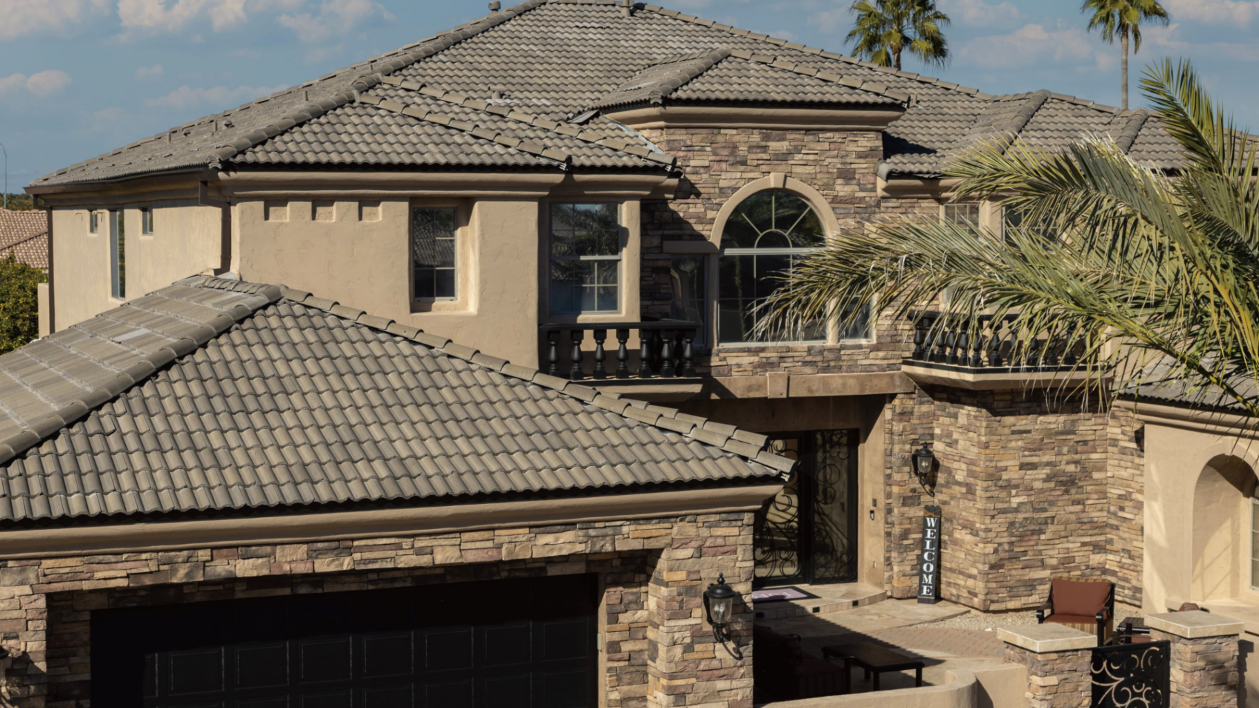The Strength and Style of Concrete Tile Roofing 2 A large, two-story house with a stone facade and stylish gray concrete tile roofing is shown. It features arched windows, a balcony, and a welcoming entrance. Palm trees are visible in the background under a partly cloudy sky.