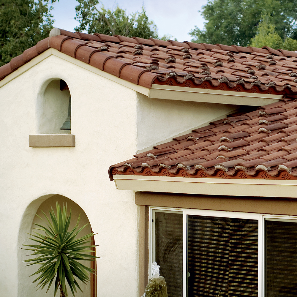 The Strength and Style of Concrete Tile Roofing 1 A house with white stucco walls features a roof covered in Boral Concrete Tiles. A small archway complements the rectangular window with blinds. In the foreground, a spiky green plant stands, while trees provide a scenic backdrop.