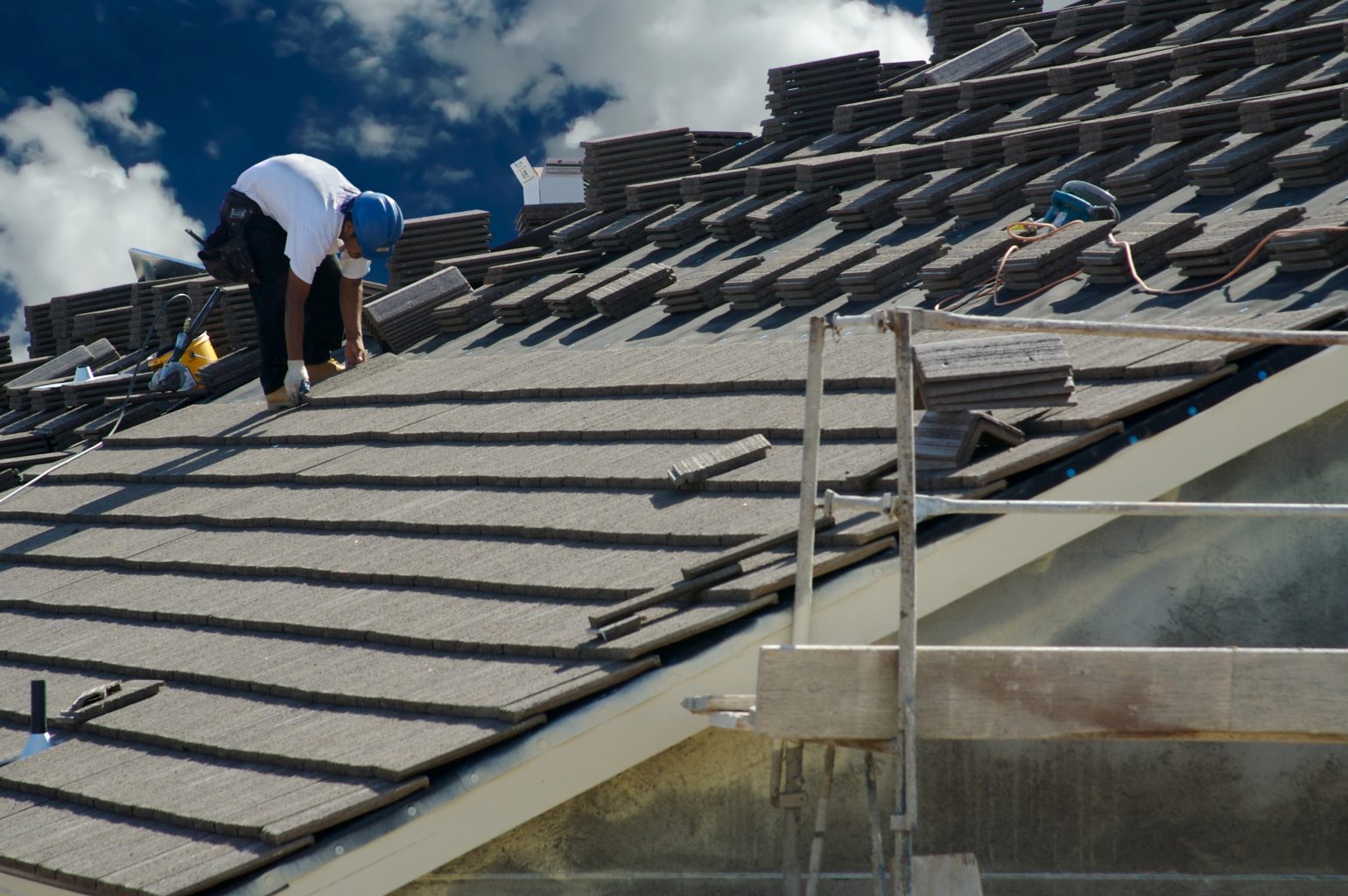 Two construction workers in helmets carefully arrange rows of dark shingles on the sloped roof, ensuring durability. With scaffolding in the foreground and a partly cloudy sky above, their work promises longevity akin to that of classic slate tile roofs.