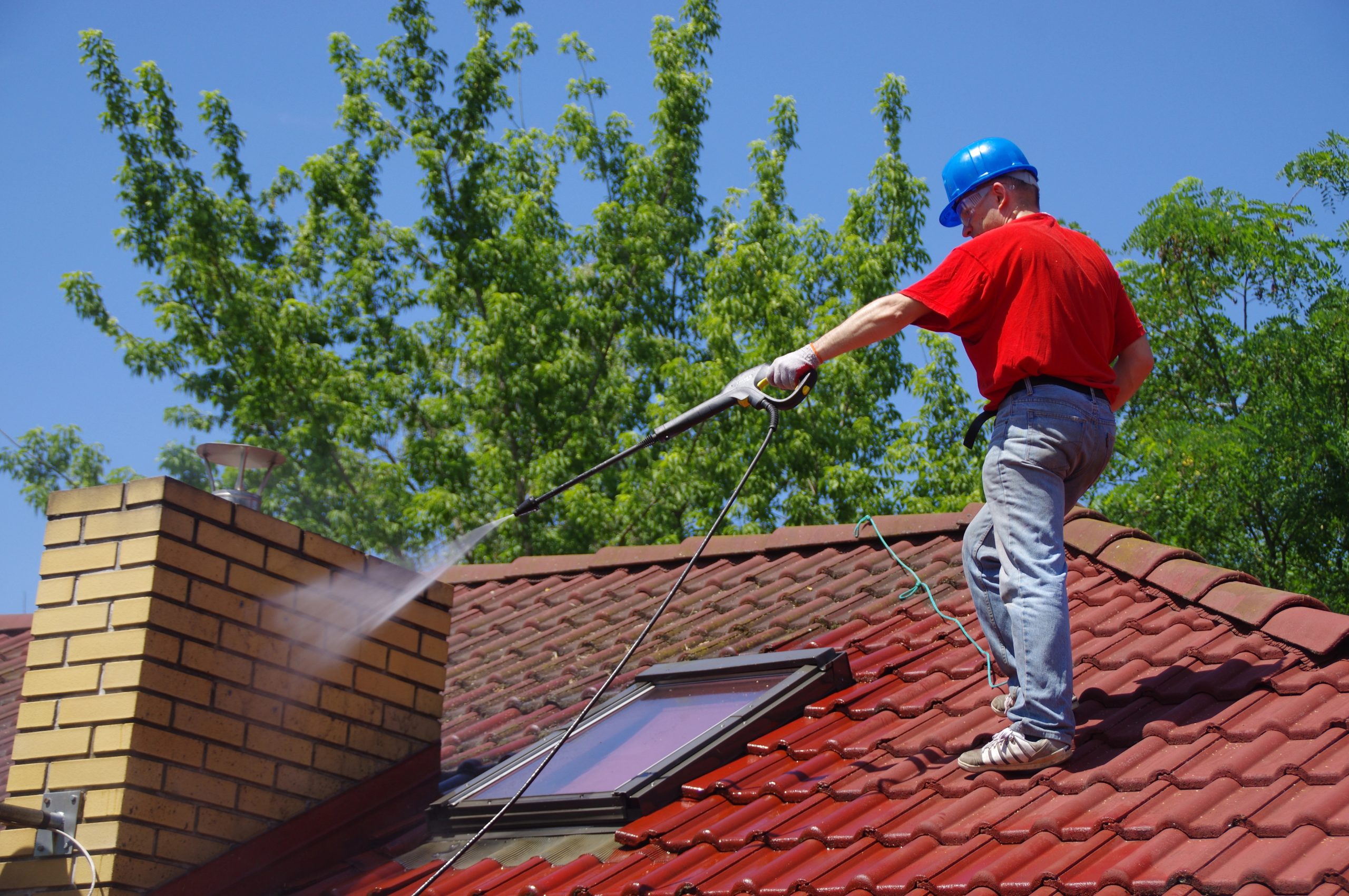 A worker in a blue hard hat and red shirt uses a pressure washer to clean a red tiled roof near a brick chimney on a sunny day, with green trees in the background.