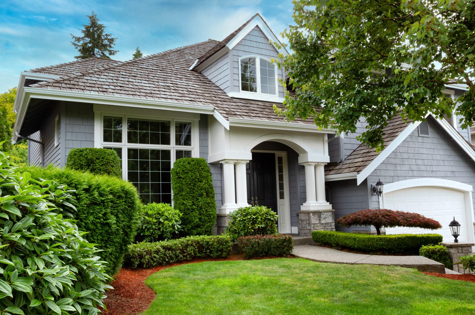 A modern grey house with white trim, large windows, and a covered entryway, surrounded by manicured shrubs, green lawn, and well-maintained landscaping under a blue sky.