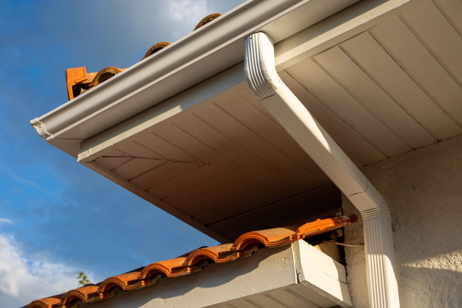 Close-up of a house roof corner with orange clay tiles and white eaves, featuring a white gutter accessory attached, set against a partly cloudy blue sky.