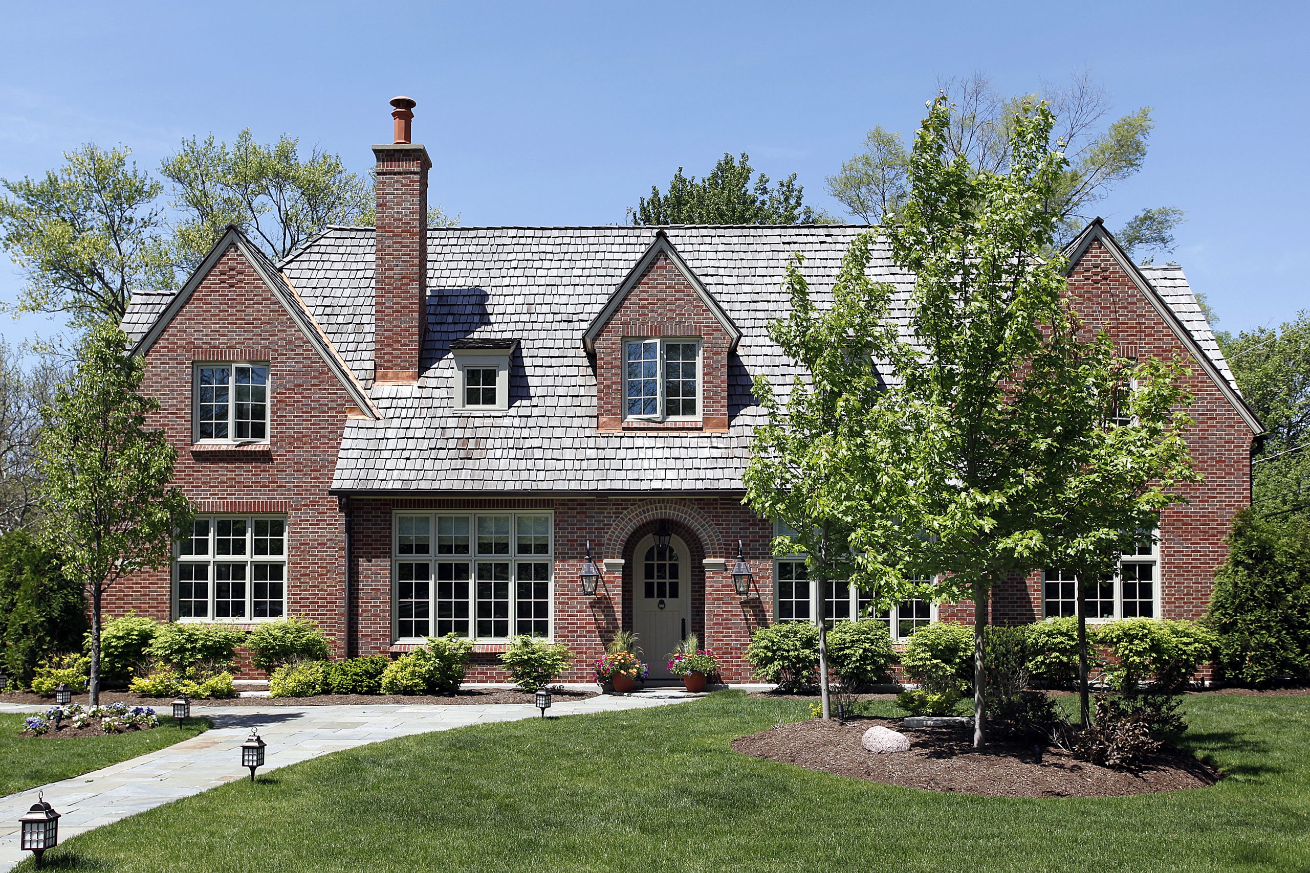 A two-story brick house with a steep, modern wood roofing, large windows, and a central arched entryway, surrounded by green lawn, trees, shrubs, and a stone walkway leading to the front door on a sunny day.