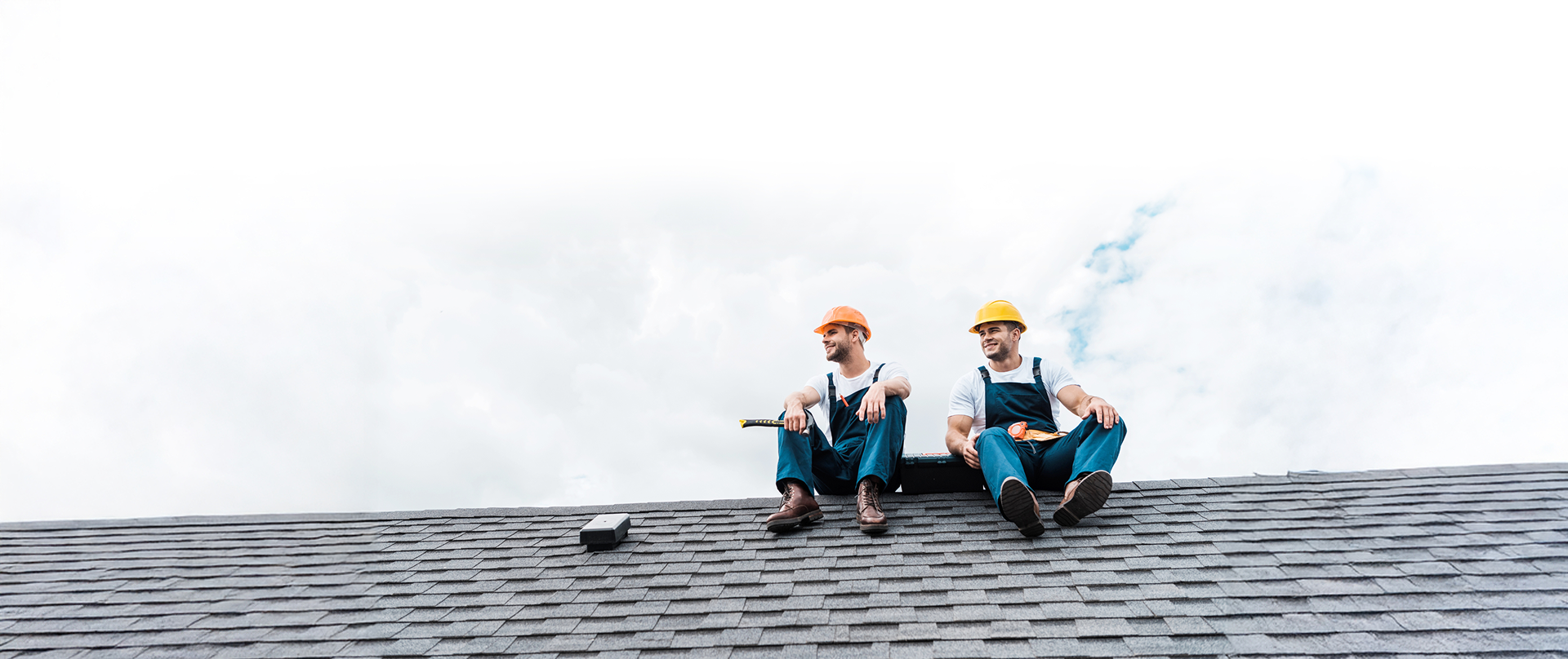 Two construction workers wearing helmets and overalls sit on a shingle roof, taking a break from a roof replacement project with tools beside them. The sky is bright and mostly cloudy in the background.