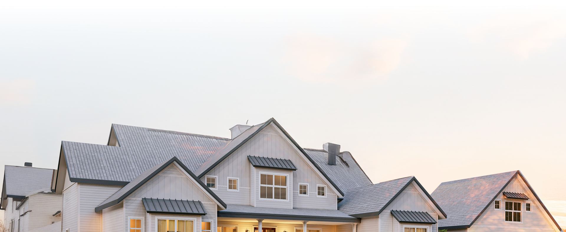 A large, modern house with gray roofs, white siding, and several peaked gables, shown in soft, evening sunlight against a pale sky.