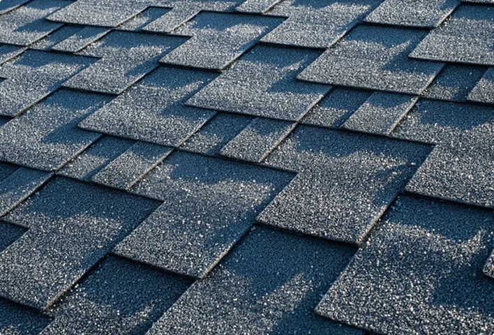 Close-up view of overlapping, textured asphalt roof shingles in a staggered pattern, with sunlight casting sharp-edged shadows across the surface.