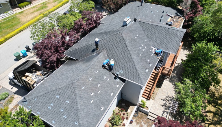 Aerial view of several workers installing new shingles on a large residential roof, with tools and materials visible. Surrounding the house are trees, a truck with debris, and a wooden deck.