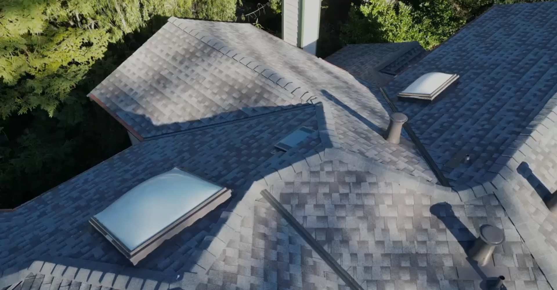 Aerial view of a house roof with gray shingles, two skylights, a chimney, and several roof vents, surrounded by green trees.
