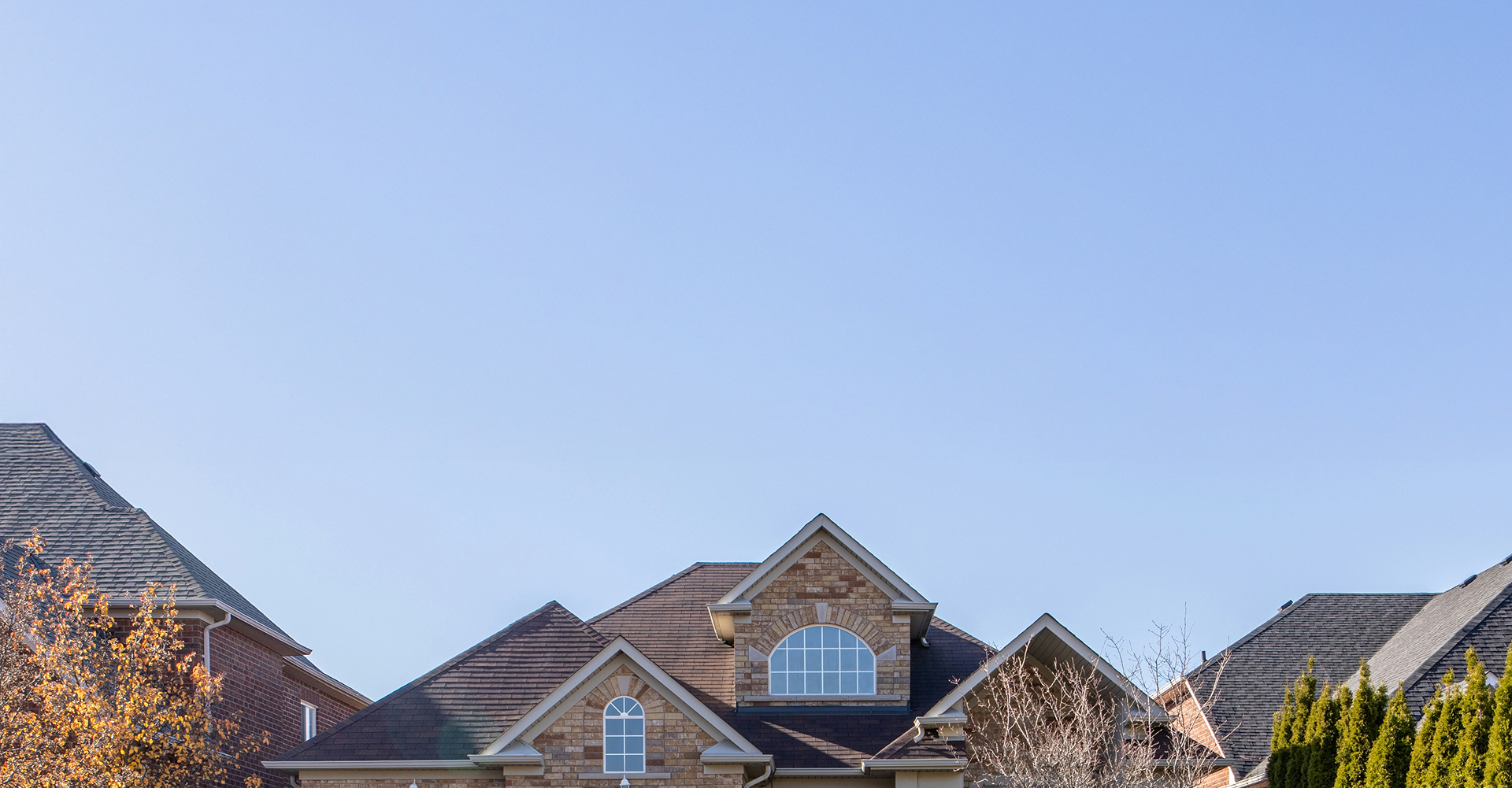 A brick house with a steep, dark roof and arched windows is framed by trees and clear blue sky, featuring new gutters from Bay 101 roofing, with parts of neighboring houses visible on either side.