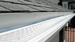 Close-up view of a house roof with gray shingles and a white gutter guard installed along the edge to prevent debris from entering the gutter.