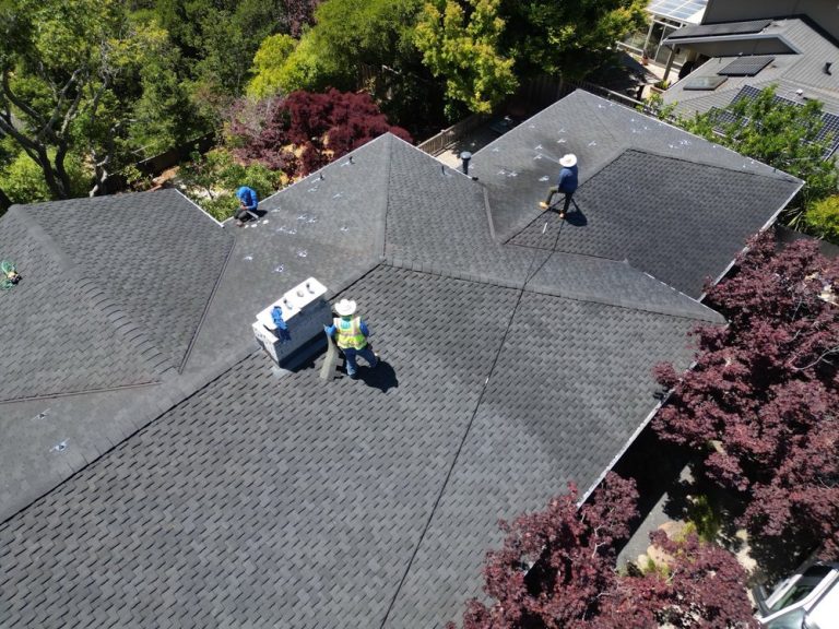 Workers wearing safety gear are installing or inspecting dark gray shingles on a large, multi-sectioned residential roof surrounded by trees and greenery on a sunny day.