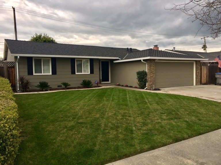 Single-story suburban house with gray siding, black shutters, a stone-accented garage, and a well-maintained green lawn under a cloudy sky. Small shrubs are planted along the front of the house.
