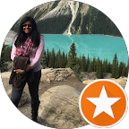 A person wearing sunglasses stands on rocky ground near a turquoise lake, with pine trees and a mountain in the background. An orange star badge appears in the corner of the image.