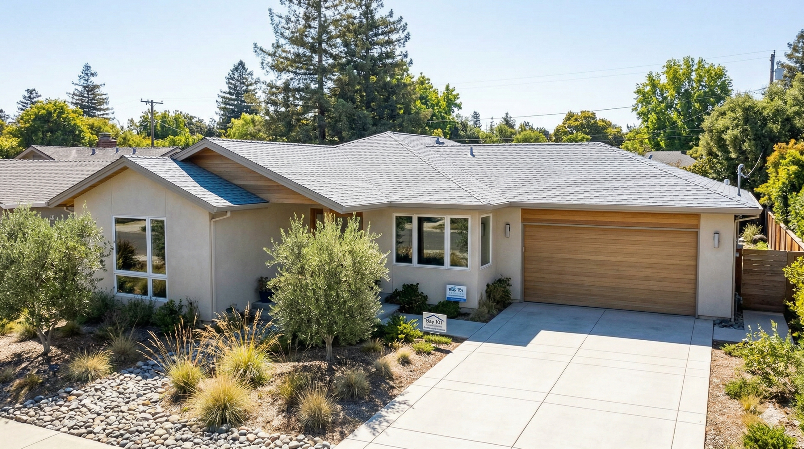 Single-story modern South Bay house with beige exterior, cool roof with stylish shingle colors, large windows, and a wooden garage door. The front yard features drought-tolerant landscaping with rocks and shrubs, plus a wide concrete driveway.